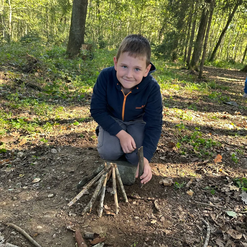 Ein junger Junge kniet auf dem Boden im Wald und lächelt, während er sich darauf vorbereitet, ein Feuer mit Stöcken zu entzünden.