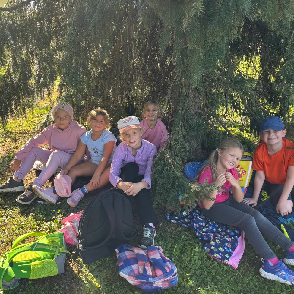 Eine Gruppe von Kindern sitzt an einem sonnigen Tag unter einem Baum. Sie lächeln und scheinen für ein Foto zu posieren.