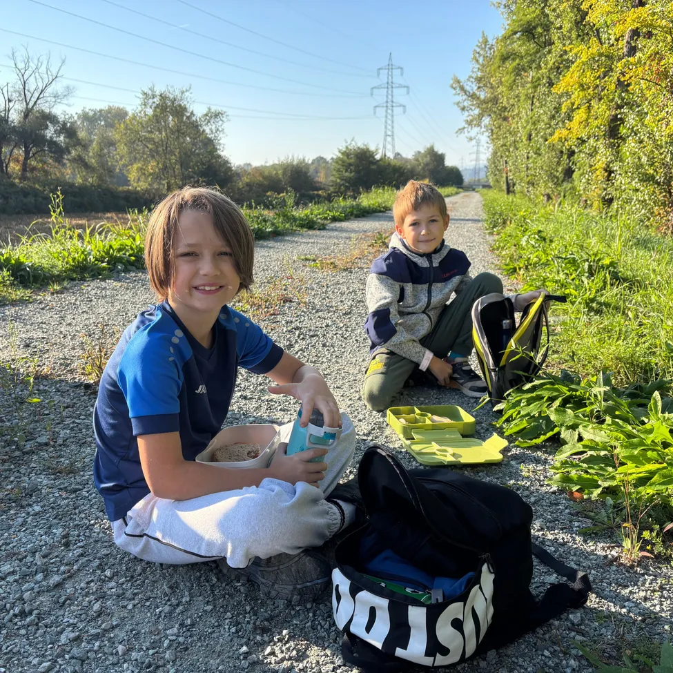 Zwei junge Jungen sitzen auf einem Kiesweg und lächeln für ein Foto. Ein Junge hält eine Schüssel und eine Flasche, während der andere Junge mit einem Rucksack und einer Brotdose sitzt.