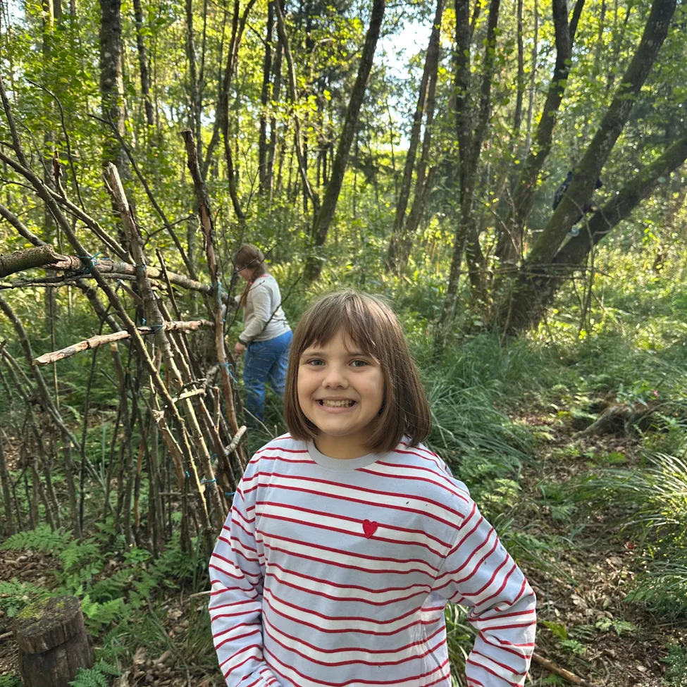 Ein junges Mädchen steht in einem Wald und lächelt, mit einem provisorischen Unterschlupf aus Ästen hinter ihr.