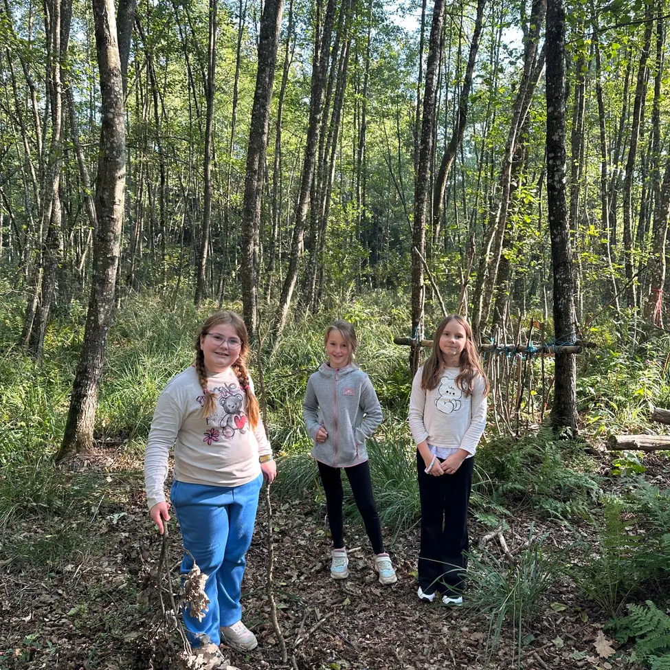 Drei junge Mädchen stehen in einem Wald. Das Mädchen auf der linken Seite hält einen Stock und lächelt. Das Mädchen in der Mitte schaut etwas an. Das Mädchen auf der rechten Seite schaut in die Kamera.