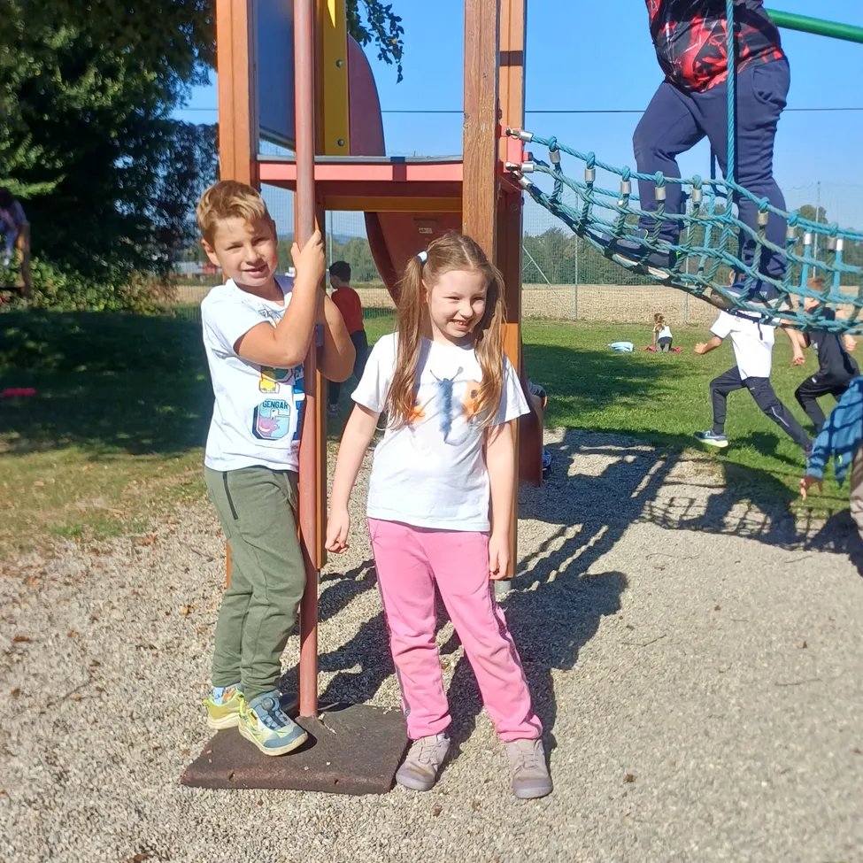 Drei Kinder spielen auf einem Spielplatz mit einer Holzstruktur, einem grünen Kletternetz und einer Rutsche.