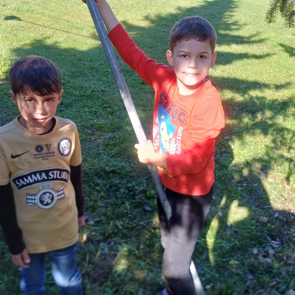 Zwei junge Jungen stehen in einem Park. Ein Junge hält einen Stock und lächelt, während der andere Junge ein gelbes Shirt mit einem Logo trägt und in die Kamera schaut.