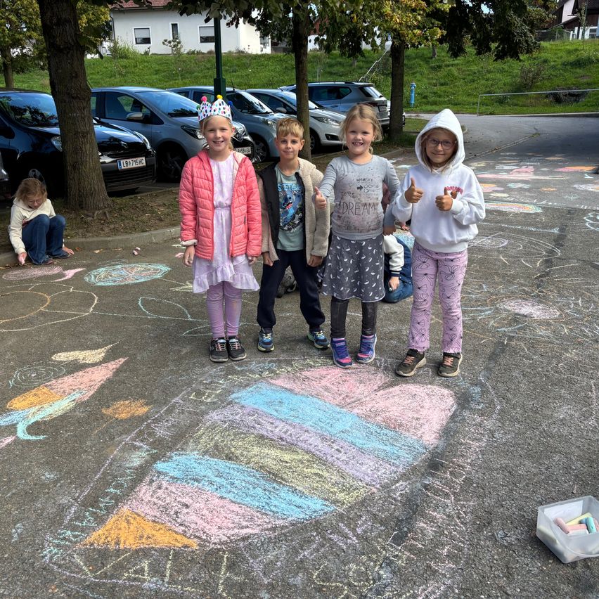 Eine Gruppe junger Kinder steht auf einem Parkplatz und posiert für ein Foto. Sie sind von bunten Kreidezeichnungen auf dem Boden umgeben.