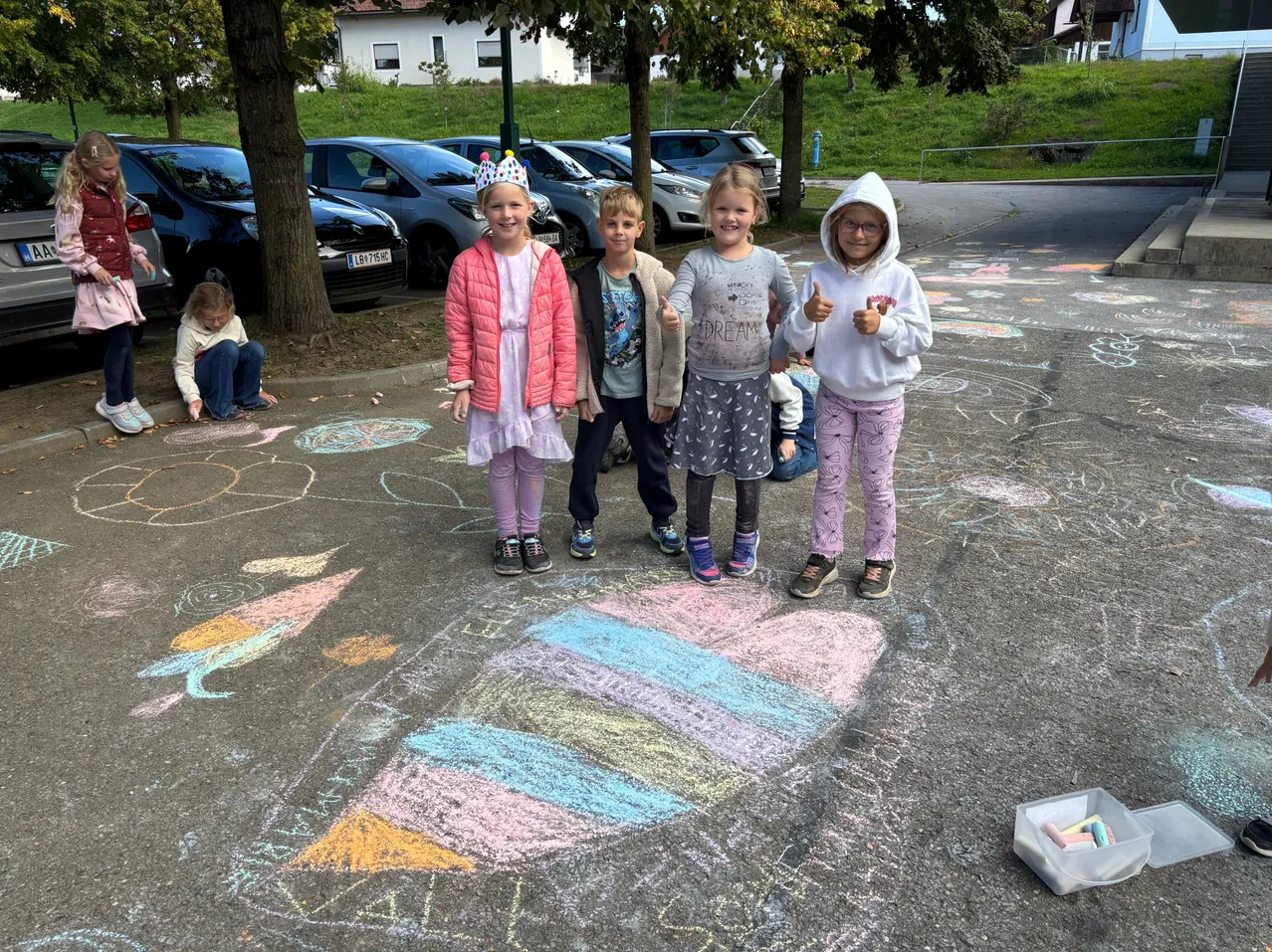 Eine Gruppe junger Kinder steht auf einem Parkplatz und posiert für ein Foto. Sie sind von bunten Kreidezeichnungen auf dem Boden umgeben.