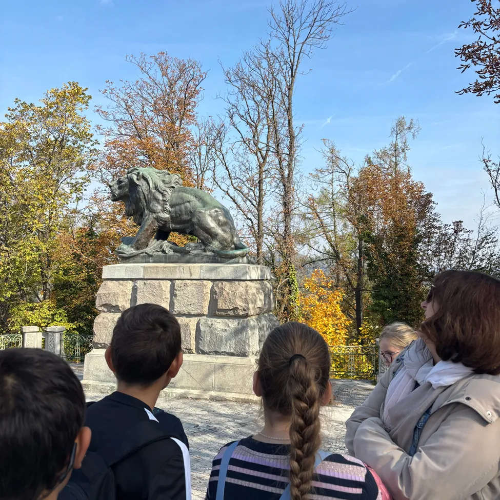 Eine Gruppe von Menschen versammelt sich um eine Löwenstatue in einem Park an einem sonnigen Tag.