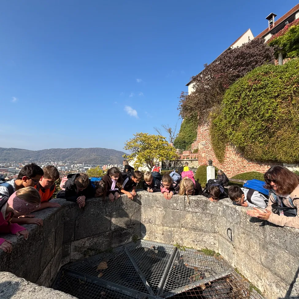 Eine Gruppe von Kindern und Erwachsenen versammelt sich um eine Steinmauer und schaut in einen Brunnen.