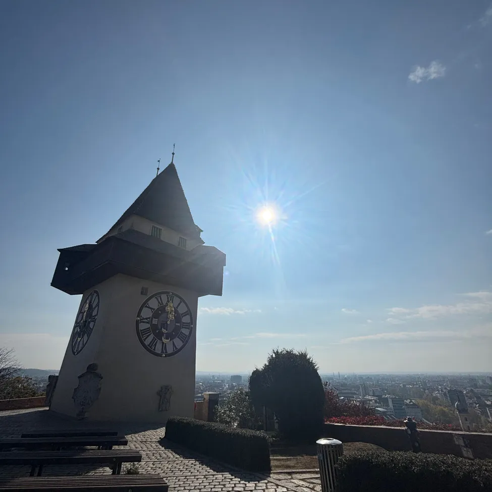 Ein Uhrturm mit zwei Uhren steht auf einem Kopfsteinpflasterweg. Der Turm hat ein spitzes Dach und ist von Grün umgeben. Die Sonne scheint hell am Himmel.