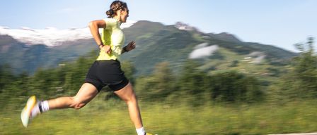 A woman in a yellow shirt and black shorts runs on a road, with a mountain range in the background, and her shadow is cast on the road.