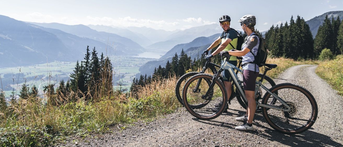 Zwei Radfahrer in Helmen und Rucksäcken stehen auf einem Schotterweg und blicken auf Berge und einen See. Sie scheinen über ihre nächste Radroute zu diskutieren.