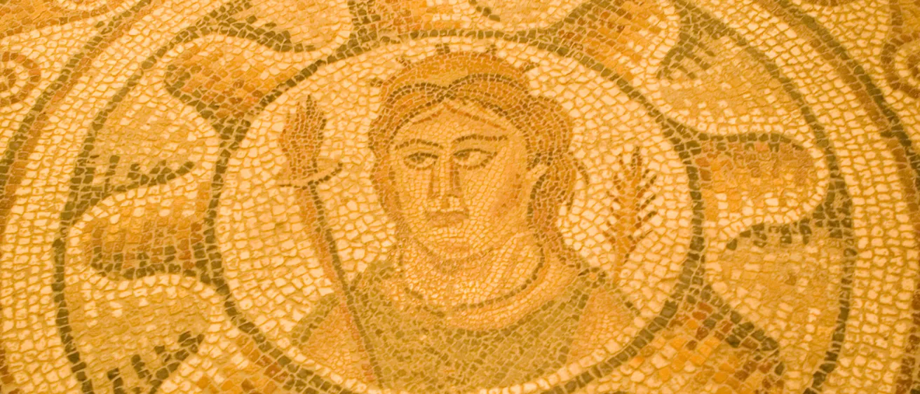 Ein Nahaufnahme eines alten Mosaikbodens zeigt ein zentrales, kreisförmiges Motiv mit einem Gesicht, umgeben von komplizierten Mustern, in warmen, erdigen Tönen.