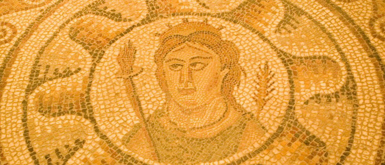 Ein Nahaufnahme eines alten Mosaikbodens zeigt ein zentrales, kreisförmiges Motiv mit einem Gesicht, umgeben von komplizierten Mustern, in warmen, erdigen Tönen.