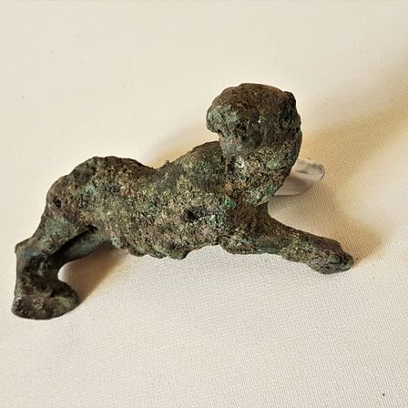 Eine antike Bronzefigur zeigt eine kauernde Gestalt, möglicherweise eine Gottheit, mit feinen Details und einer grünlichen Patina. Sie ruht auf einer weißen Oberfläche.