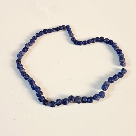 Eine Halskette aus blauen Perlen wird auf einer weißen Oberfläche in Herzform angeordnet präsentiert. Die Perlen variieren in Größe und Form.