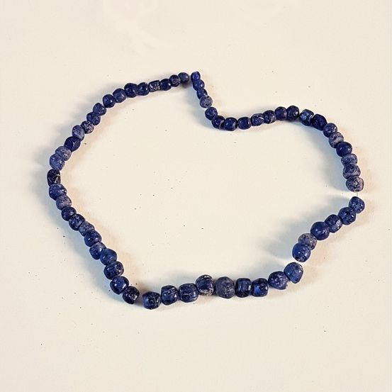Eine Halskette aus blauen Perlen wird auf einer weißen Oberfläche in Herzform angeordnet präsentiert. Die Perlen variieren in Größe und Form.