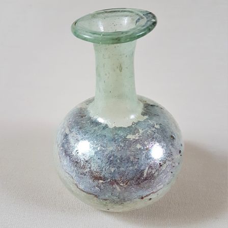 Eine antike Glasvase mit grünlich-blauer Farbe, einem schmalen Hals und einem kugelförmigen Körper, die auf einer weißen Oberfläche ruht.