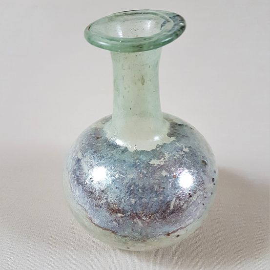 Eine antike Glasvase mit grünlich-blauer Farbe, einem schmalen Hals und einem kugelförmigen Körper, die auf einer weißen Oberfläche ruht.