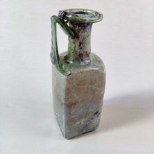 Ein alter grüner Glaskrug mit einem Griff steht auf einer weißen Oberfläche. Er hat einen rechteckigen Körper und eine runde Öffnung oben.