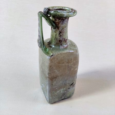 Ein alter grüner Glaskrug mit einem Griff steht auf einer weißen Oberfläche. Er hat einen rechteckigen Körper und eine runde Öffnung oben.