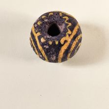 Eine kleine, runde, schwarze Perle mit gelben Mustern liegt auf einer weißen Oberfläche. Die Perle hat ein Loch in der Mitte und unebenen Oberflächen.