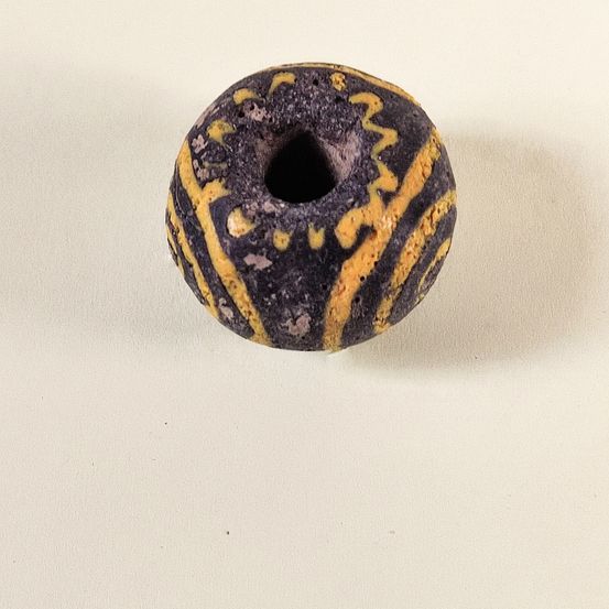 Eine kleine, runde, schwarze Perle mit gelben Mustern liegt auf einer weißen Oberfläche. Die Perle hat ein Loch in der Mitte und unebenen Oberflächen.