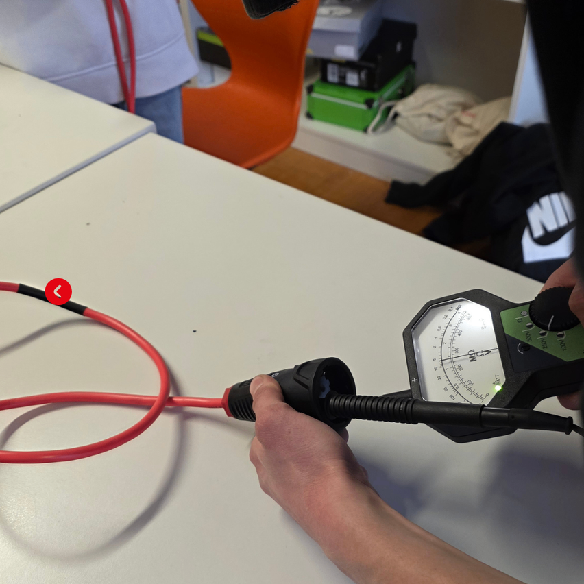 Eine Person benutzt ein schwarzes Werkzeug mit einer Anzeige darauf, das an ein rotes Kabel angeschlossen ist. Ein oranger Stuhl und ein weißer Schreibtisch sind im Hintergrund.