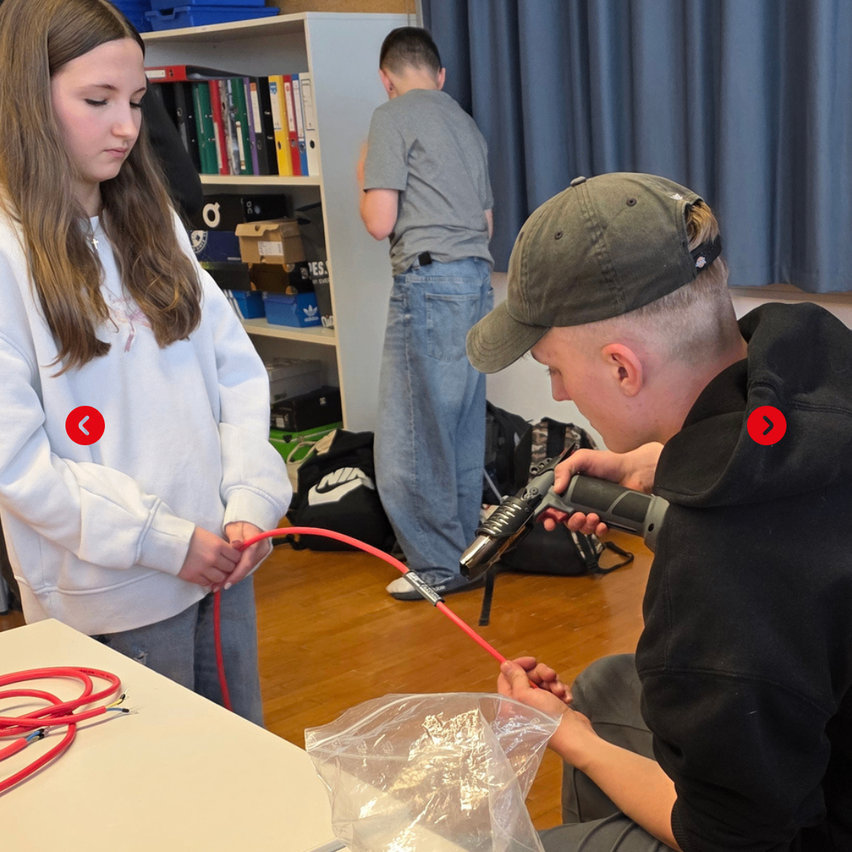 Drei Schüler in einem Klassenzimmer, einer benutzt ein Werkzeug an einem roten Kabel, ein anderer hält es, und ein dritter beobachtet. Regale mit Büchern und Taschen im Hintergrund.