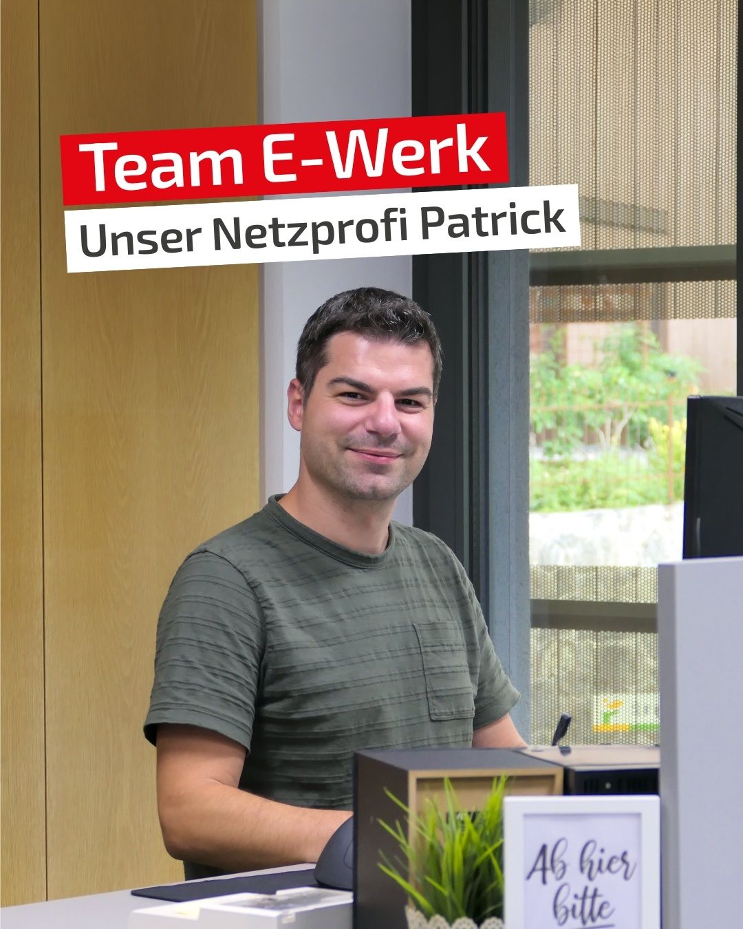 Ein Mann in einem grünen T-Shirt lächelt in die Kamera und steht vor einem Schreibtisch mit einer Pflanze und einem Computermonitor. Im Hintergrund ist ein Fenster mit Blick auf Bäume und ein rotes Schild zu sehen, das 'Team E-Werk Unser Netzprofi Patrick' besagt.