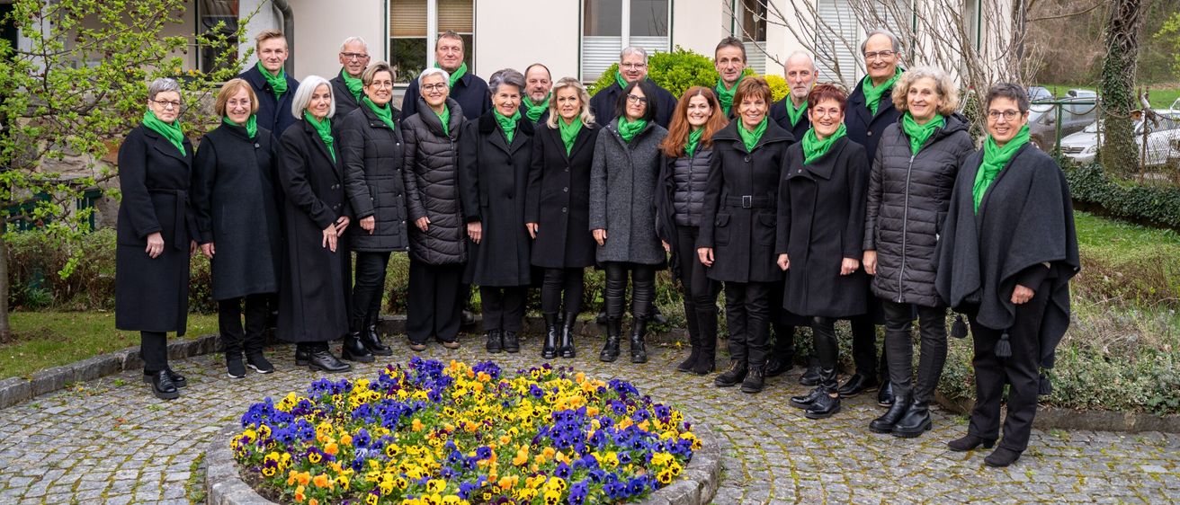 Ein Chor in grünen Schals steht vor einem Gebäude mit einem Blumenbeet im Vordergrund.