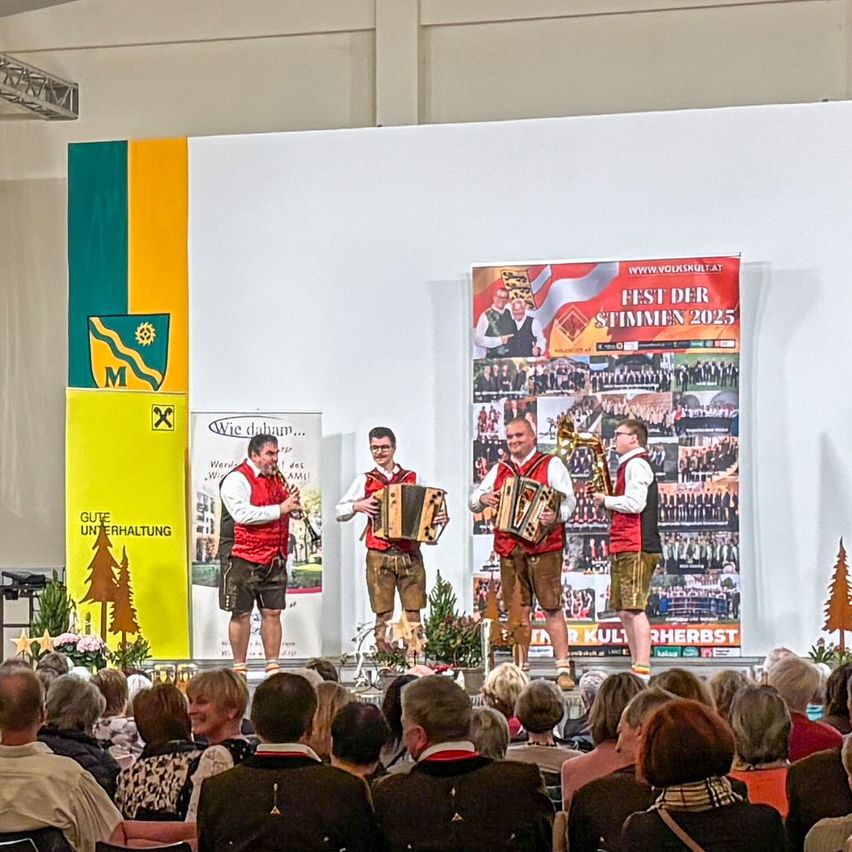 Drei Männer spielen auf der Bühne bei einer kulturellen Veranstaltung. Sie spielen Klarinette und Akkordeons, mit einem Banner hinter ihnen, das 'Fest der Stimmen 2025' steht. Ein Publikum sitzt vor ihnen.