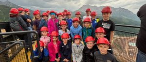 Eine Gruppe von Kindern in roten Helmen posiert für ein Foto auf einer gelben Plattform, mit einem bewölkten Himmel und Bergen im Hintergrund.