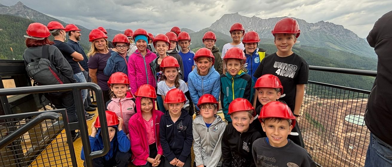 Eine Gruppe von Kindern in roten Helmen posiert für ein Foto auf einer gelben Plattform, mit einem bewölkten Himmel und Bergen im Hintergrund.