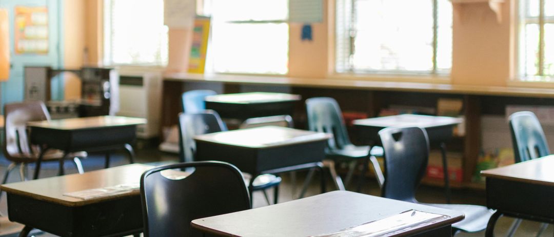 Ein leerer Klassenraum mit mehreren Schreibtischen und Stühlen, einer Uhr an der Wand und einer Tafel mit einigen Papieren.