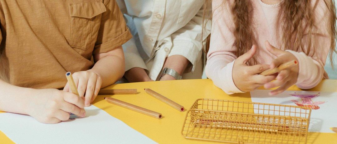 Eine Frau und zwei Kinder arbeiten gemeinsam an einem Tisch. Die Frau schaut auf die Kinder hinunter. Der Junge hält einen Bleistift und das Mädchen einen Stock. Sie scheinen ein Artprojekt zu machen.