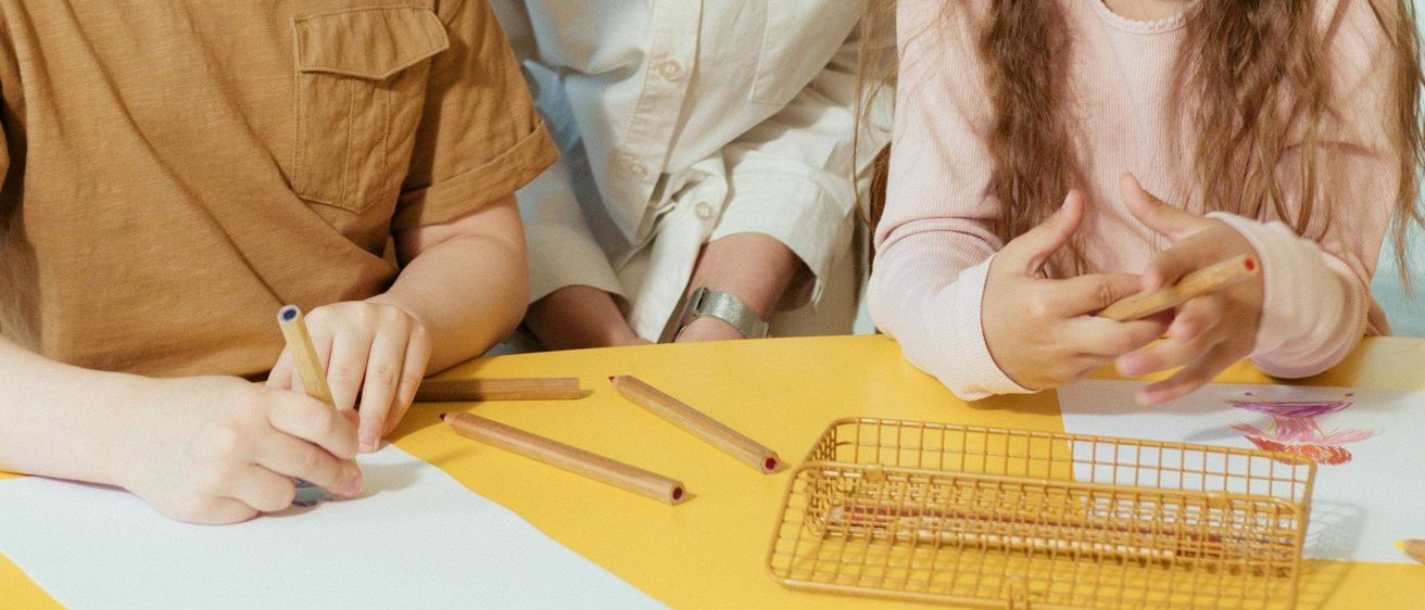 Eine Frau und zwei Kinder arbeiten gemeinsam an einem Tisch. Die Frau schaut auf die Kinder hinunter. Der Junge hält einen Bleistift und das Mädchen einen Stock. Sie scheinen ein Artprojekt zu machen.