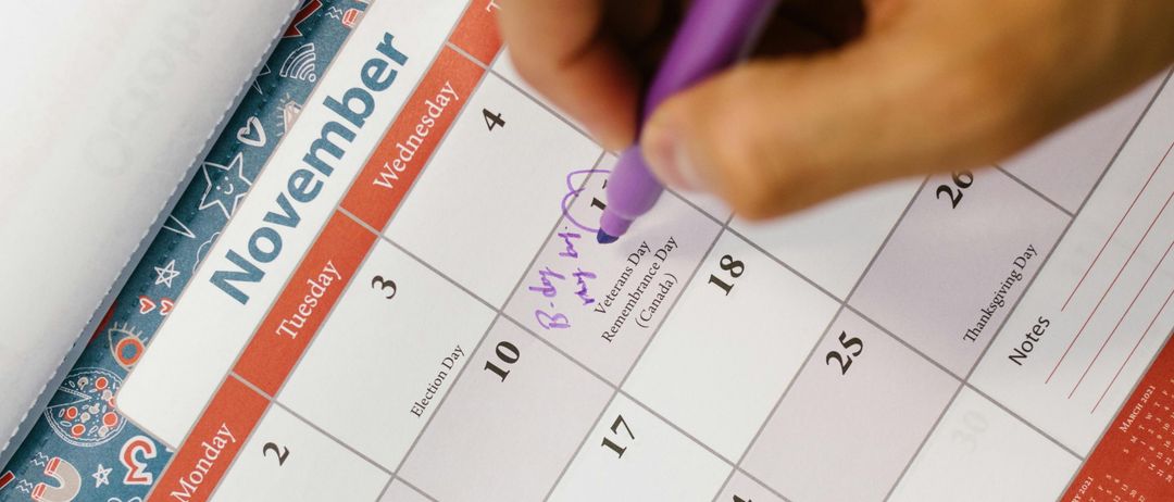 Eine Hand mit einem lila Stift schreibt auf einen Kalender und markiert den Veterans Day. Der Kalender hat Daten vom 1. November bis zum 30. November.