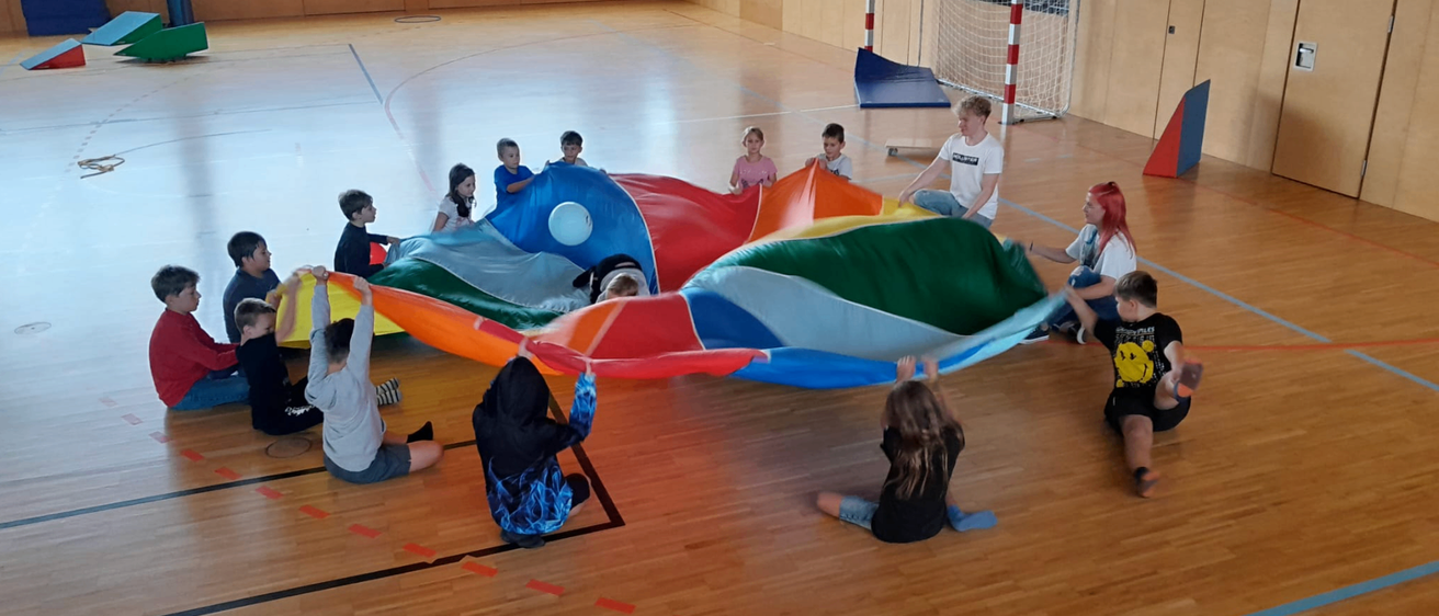 Mehrere Kinder spielen auf einem großen, bunten Luftkissen in einer Turnhalle, mit einem weißen Ball oben drauf. Ein Kind hält eine Ecke.