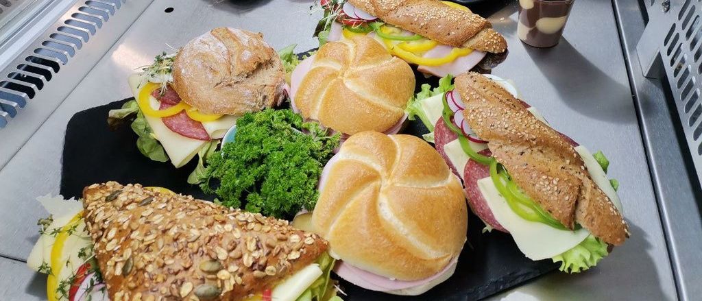 Eine Auswahl an Sandwiches, Brötchen und Salaten ist auf schwarzen Tabletts ausgestellt. Einige Sandwiches haben Fleisch und Gemüse, während andere nur ein Stück Fleisch haben. Es gibt auch Äpfel und einen Becher Getränk.