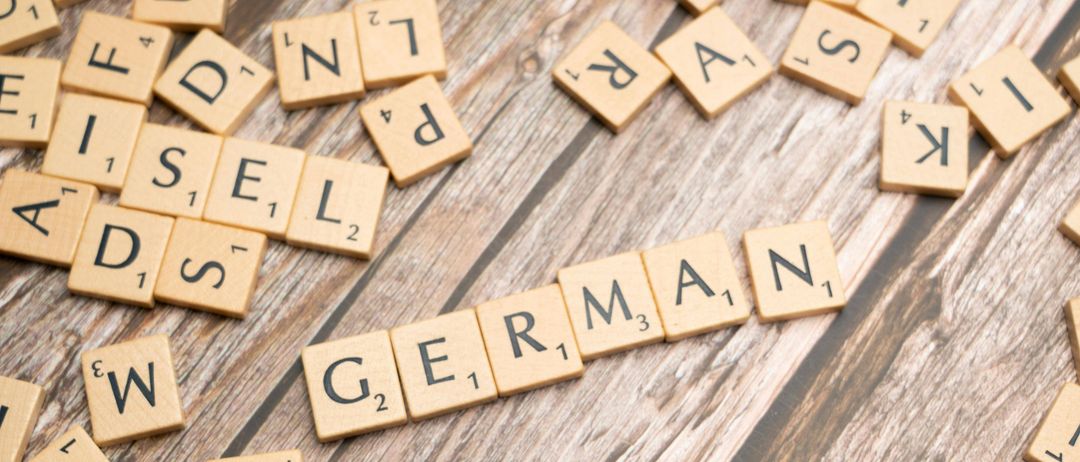 Holzne Scrabble-Steine bilden das Wort German auf einem Holztisch. Jeder Stein hat einen Buchstaben, einige haben auch Zahlen.