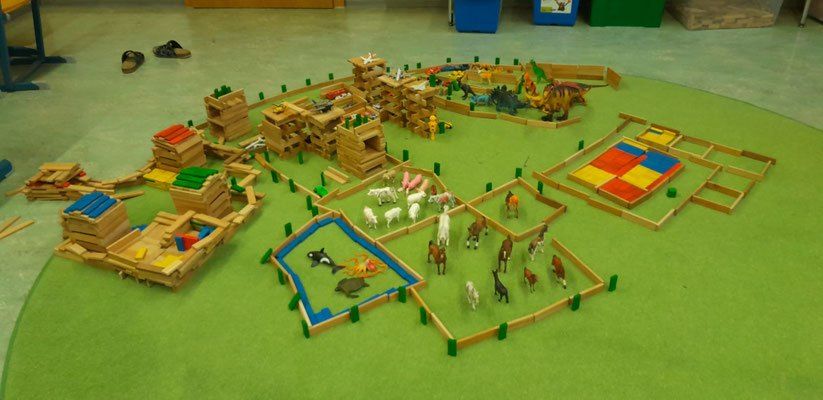 Eine Miniaturwelt aus Holzklötzen und Figuren ist auf einem grünen Teppich ausgelegt. Verschiedene Strukturen und Tiere sind enthalten.