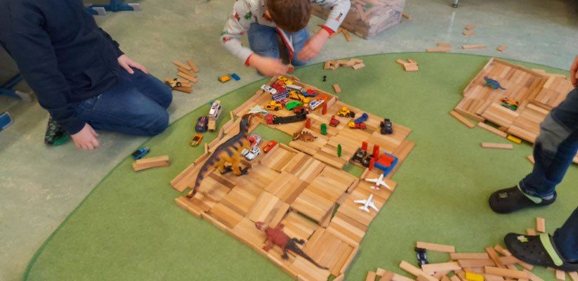 Zwei Kinder spielen mit einem großen Holzbaustein-Set auf einem grünen Teppich. Ein Kind ordnet Autos und Dinosaurier an, während das andere neben ihnen sitzt. Verschiedene Spielzeuge und Blöcke sind herum verstreut.
