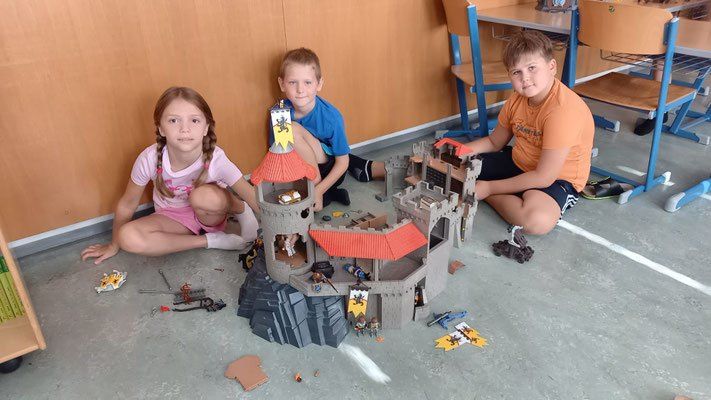 Drei Kinder spielen mit einem Burgspielzeug auf dem Boden, ein Mädchen und zwei Jungen. Sie sitzen um die Burg mit rotem Dach und kleinen Figuren herum.