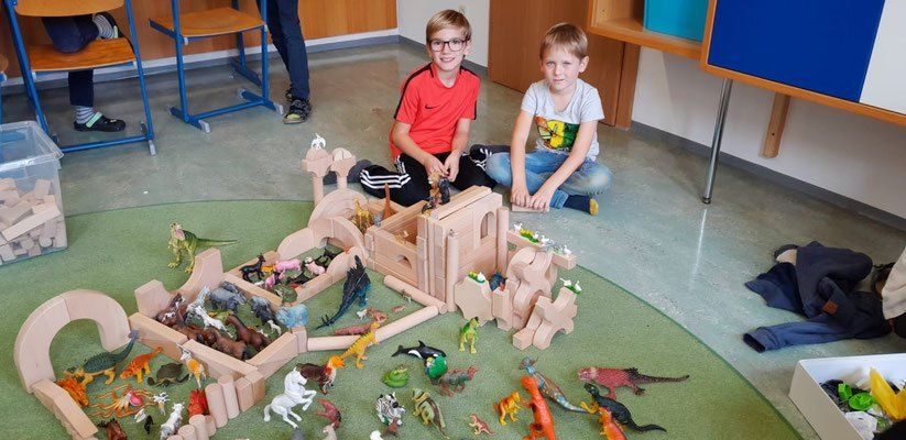Zwei Kinder spielen mit Holzspielzeug, darunter Dinosaurier und andere Tiere, auf einem grünen Teppich in einem Klassenzimmer.