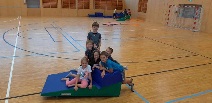 Mehrere Kinder in einer Turnhalle, posieren für ein Foto, während sie auf einer blauen und grünen Matte auf einer Rampe sitzen.