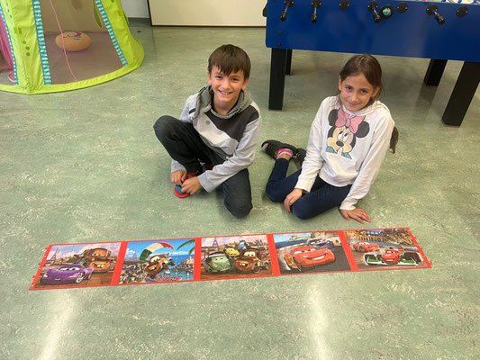 Zwei Kinder sitzen lächelnd auf dem Boden mit einem Puzzle-Teppich von Cars-Charakteren vor ihnen. Ein Spielhaus und ein Tischfußball sind im Hintergrund.