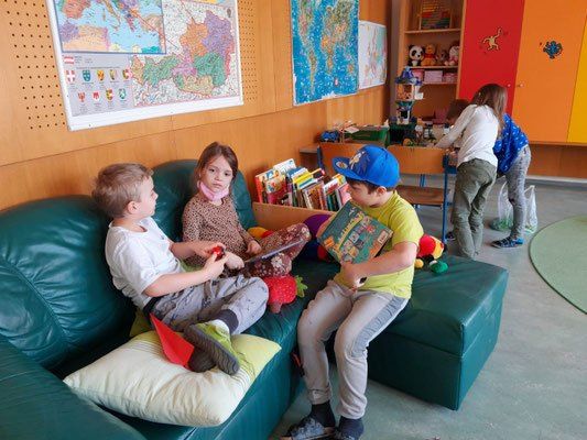 Vier Kinder sitzen auf einer grünen Couch in einem Klassenzimmer, umgeben von Spielzeug und Büchern. Ein Kind hält ein Buch, ein anderes spielt mit einem Tablet.
