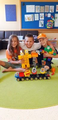 Drei Kinder spielen mit Spielzeugzügen auf einem grünen Teppich. Ein Junge hält ein Spielzeug in den Händen. Der Raum hat eine Couch und einen Anschlagkasten.