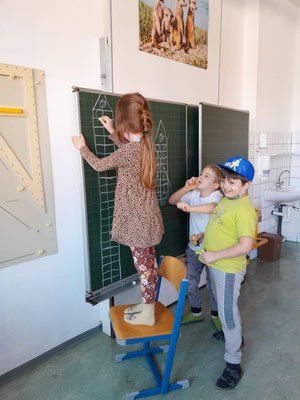 Ein Mädchen steht auf einem Stuhl und schreibt auf eine Tafel, während zwei Jungen zuschauen. Sie befinden sich in einem Klassenzimmer mit einem Waschbecken und einem Bild an der Wand.
