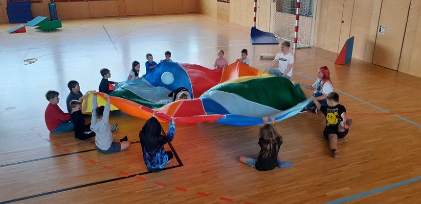 Eine Gruppe von Kindern spielt mit einem großen, farbenfrohen Fallschirm in einer Turnhalle. Einige sitzen auf dem Boden, während andere stehen und die Kanten des Fallschirms halten. Ein Torpfosten und einige Holzplatten sind im Hintergrund.