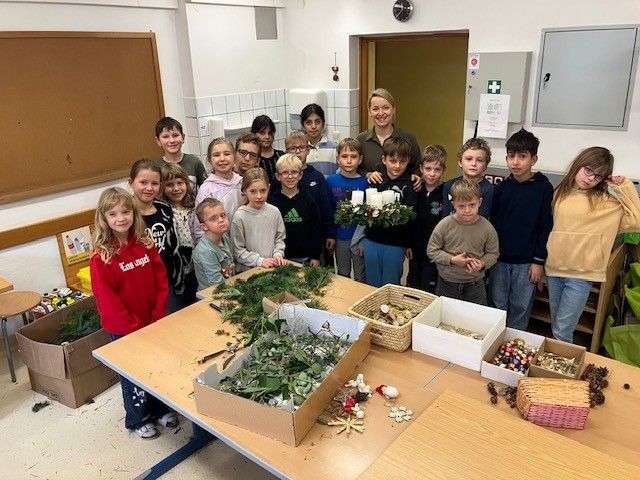 Eine Gruppe von Kindern und einer Frau befindet sich in einem Raum mit einem Tisch voller Materialien. Sie lächeln und scheinen etwas zu basteln.